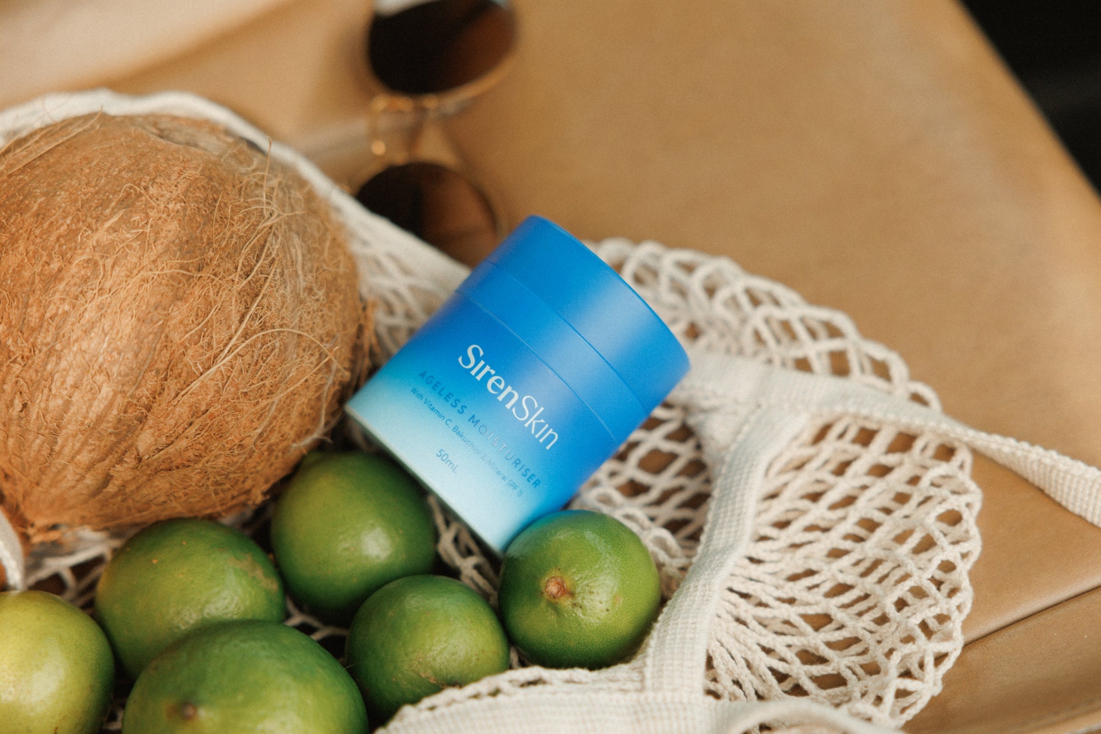 Blue skincare product bottle in a mesh bag with coconuts and limes.
