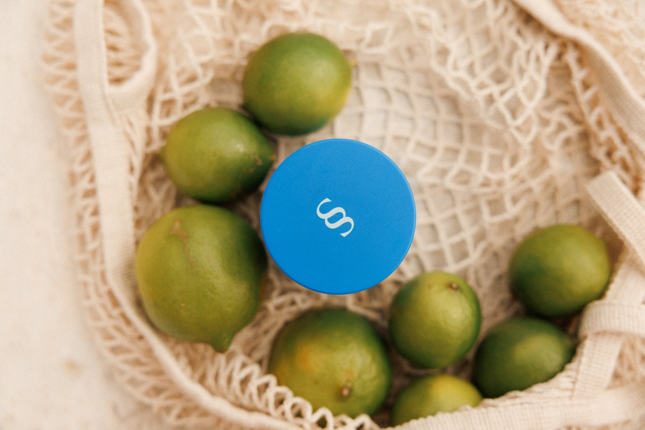 A blue bottle of Siren Skin Moisturiser placed on a surface with green limes and white meshed bag