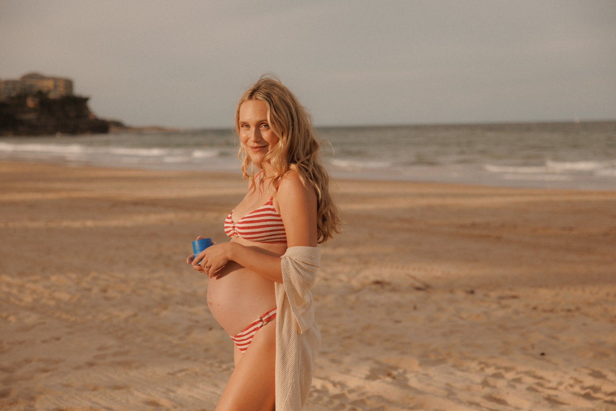 Woman in a striped swimsuit standing on a beach with ocean waves in the background and skincare in hand